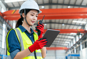 Female industrial engineer using a tablet and walkie-talkie in a factory. Asian woman in a hard hat and safety vest at a manufacturing plant. Communication and technology in heavy industry