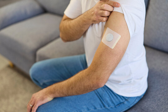 Man shows arm patch for diabetes sensor. The glucose monitor is a wearable medical device for health tracking at home, secured with adhesive on the arm. Healthcare monitoring and diabetic control.