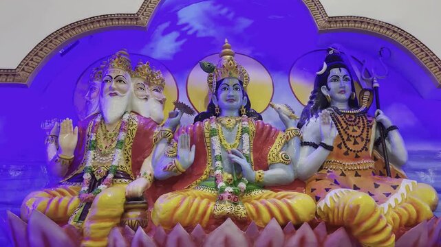 Hindu Trimurti&mdash;Lord Brahma, Lord Vishnu, and Lord Shiva&mdash;seated together in divine harmony inside a temple shrine. The colorful idols are adorned with ornate crowns, jewelry, and symbolic hand gestures