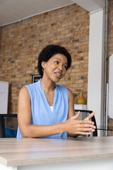 Naklejka premium African American woman in blue blouse sitting at table in modern workspace, speaking by whiteboard