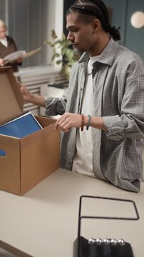 Vertical of gen Z African American male employee packing his office supplies and personal items into cardboard box and leaving office after being fired