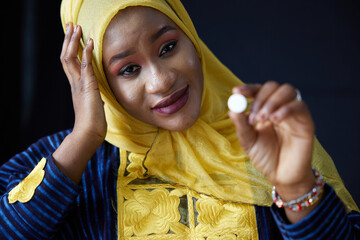 Muslim woman thoughtfully examining a single pill, wearing a yellow headscarf, with a look of concern on black background