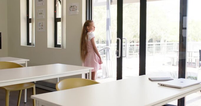 Preteen girl in gingham dress walking from glass doors to table with laptop, notebook, copy space