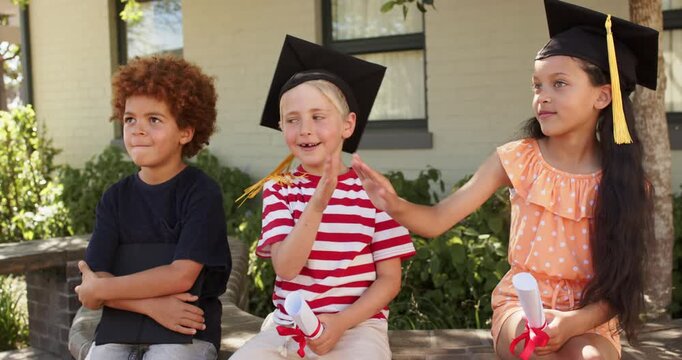 Schoolchildren on bench, middle child lifting hand starting high-five celebrating caps, diplomas