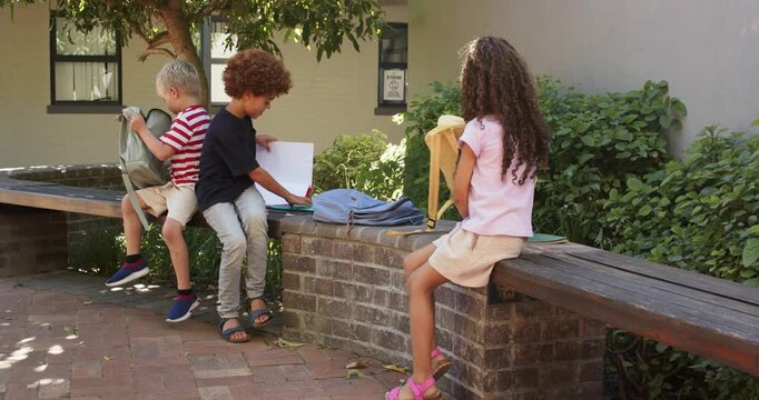 Three schoolchildren sitting on wall at school during break, sorting backpacks, opening red binder