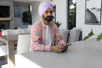 Naklejka premium Indian man wearing lavender turban sitting at gray kitchen island holding smartphone in dining area