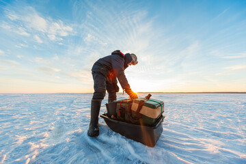 A lone angler in heavy winter gear readies a sled with tackle on a vast frozen lake at sunrise. Low sun flares, long shadows, wind shaped ice, wide and low shot.