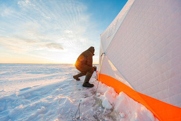A bundled angler kneels by a drilled hole, securing a line near a quilted white and orange pop up...