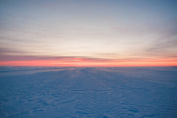 Fototapeta premium A wide frozen expanse shows faint tracks and distant huts for ice fishing. Low sun casts long shadows toward the horizon. Soft light and minimalist composition suggest isolation.