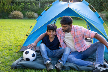 South asian father and son sitting on pad in front of blue tent with soccer ball