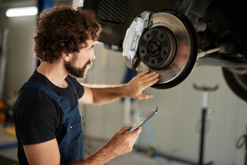 A car mechanic closely examines the brake components of a vehicle while using a clipboard in a well-lit workshop. The mechanic is focused and dedicated to quality repairs.