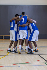 Diverse male basketball teammates huddling in circle on wood court wearing jerseys
