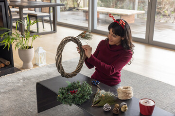 Naklejka premium Woman wearing sweater sitting on home floor assembling wreath with vine base, twine, copy space