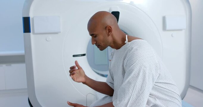 African American man in gown sitting exam table by scanner, cupping face, preparing for scan
