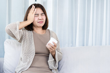 Asian middle-aged woman suffering from  headache or migraine while holding a white medicine bottle on a sofa. Healthcare concept of self-medication, stress, and pain relief at home