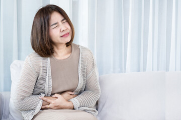 Mature Asian woman sitting on sofa suffering from severe stomach ache and abdominal pain. Healthcare concept of gastritis, food poisoning, or menstrual cramps with negative facial expression
