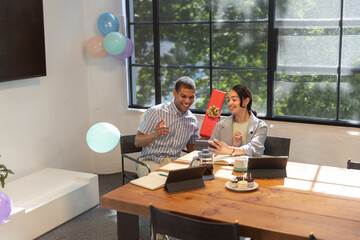 Diverse coworkers exchanging red gift and using smartphone at meeting table, pastries, balloons