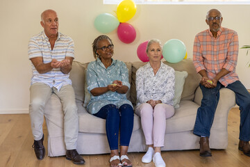 Senior friends sitting on beige couch in living room holding confetti near plant, pastel balloons © wavebreak3