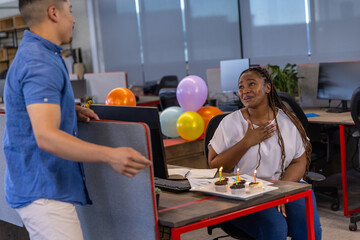 Diverse coworkers sharing birthday surprise with tray of cupcakes, balloons at desk, copy space
