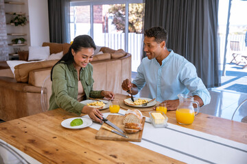 Diverse couple eating breakfast at home dining table with scrambled eggs, orange juice