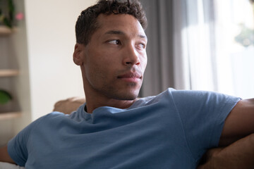 Man reclining on sofa and gazing toward window with sheer curtains, potted plant in living room