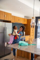 Mother lifting daughter laughing at marble countertop island in kitchen with backpack