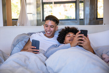Diverse male partners reclining side by side on bed under duvet using smartphones in bedroom