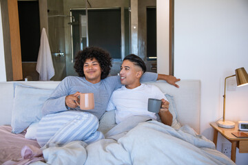 Obraz premium Diverse men wearing pajamas, leaning against headboard, sipping coffee from mugs in master bedroom