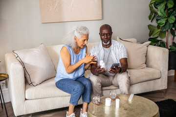 Diverse senior couple reviewing medication instructions on smartphone and pill bottles on home sofa