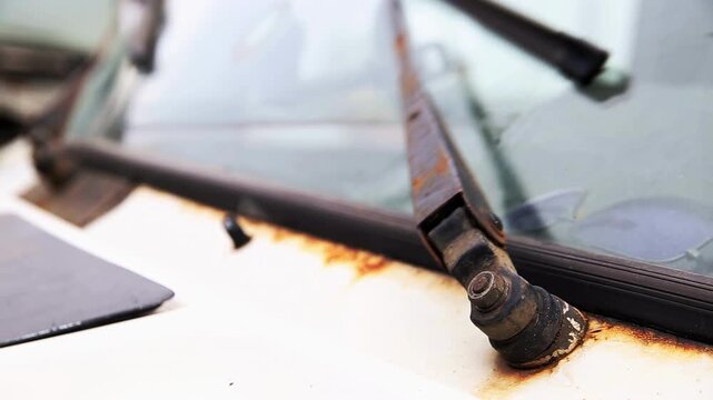 Severely rusted windshield wiper on abandoned white truck. Corrosion on metal arm and base. Blurry interior reflection. Image depicts decay, aging, and neglect of vehicle.