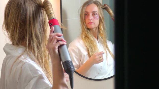 Woman curling hair with electric styler while looking in bathroom mirror