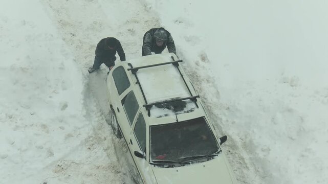 Two people push white car stuck in heavy snow on unplowed road. Winter weather creates hazardous conditions for transport as people work to free vehicle.