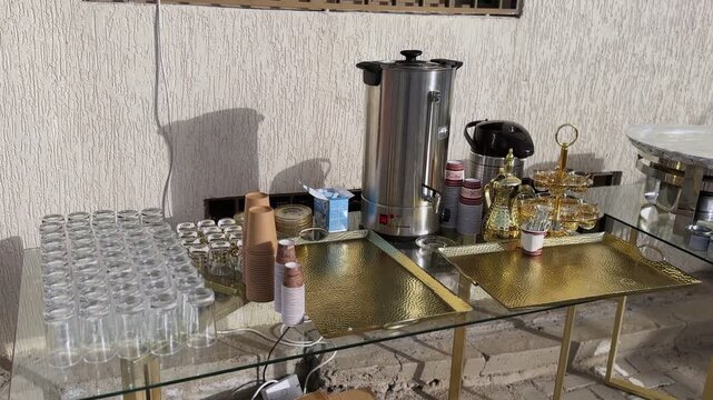 Arabic coffee and tea service setup with hot water urn, dallah pot, glass tea cups, disposable cups and golden trays on buffet table, indoor event hospitality station.