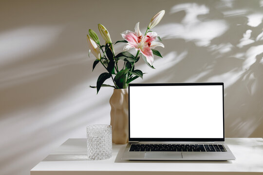 Elegant laptop mockup with white screen on white table and vase with flower with glass of water, aesthetic leaves shadows. Laptop for web site promotion, blog, design,home office or branding