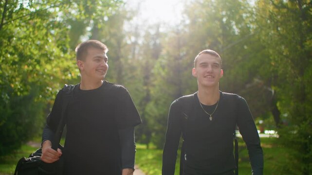 two friends walking under sunlit trees laughing and talking returning from training casual duffel bag and relaxed stride warm backlight candid smiles joyful camaraderie weekend energy