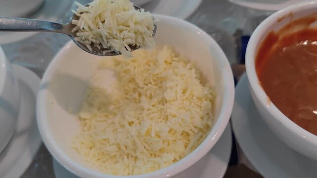 Close-up shot of freshly grated cheese held on a spoon above a white bowl filled with shredded cheese. A bowl of tomato sauce is visible beside it, creating a cooking and food preparation concept. Per