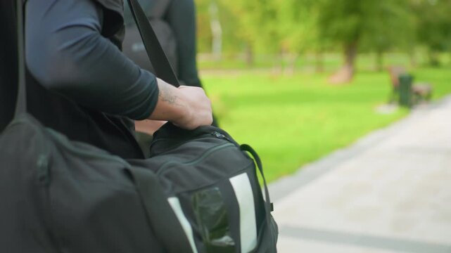 closeup white man with bag, firm grip on strap and shoulder as he adjusts duffel, black casual top, park pathway and green trees in background, mood of readiness and anticipation, roles implied