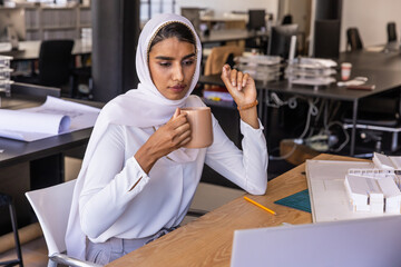 Obraz premium Indian woman sitting at design studio desk holding coffee mug while reviewing laptop with models
