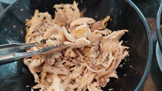 Close-up view of freshly shredded cooked chicken in a black container, held with metal tongs. Detailed texture of tender chicken meat, perfect for concepts related to food preparation, cooking process