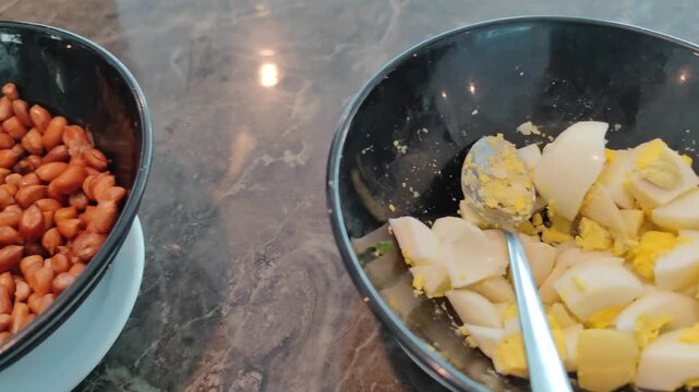 Video shows two black bowls placed on a marble surface. One bowl contains cooked brown beans, while the other holds chopped boiled eggs with visible yolks and whites. A metal spoon rests inside the eg