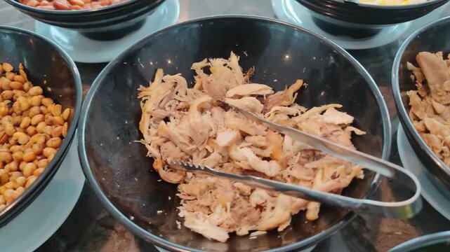Close-up view of freshly shredded cooked chicken in a black container, held with metal tongs. Detailed texture of tender chicken meat, perfect for concepts related to food preparation, cooking process