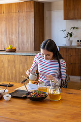 Sauce streaming from small white pitcher into noodle bowl on dining table with salad and pitcher