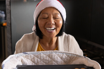 Naklejka premium Senior Asian woman leaning over baby bassinet with quilted blanket in living room wearing Santa hat
