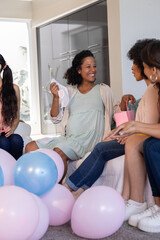 Diverse female friends sitting in living room at home examining baby bodysuit, pastel balloons