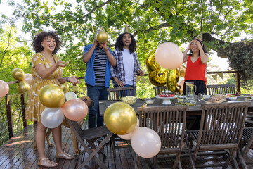 Diverse friends celebrating birthday on wooden deck, cake, balloons, lemonade, small brown dog