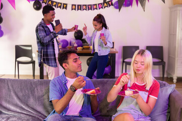 Obraz premium Diverse friends celebrating birthday on grey couch with banner balloons holding cake, pink cups