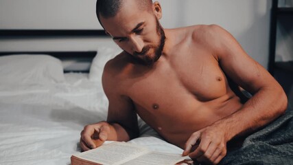 An attractive man with an athletic body reads a book in bed before going to sleep.