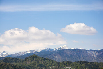 A wide landscape view of the distant snow capped peaks of the Julian Alps rising above the lush green forests and hills of the Gorenjska region in Slovenia
