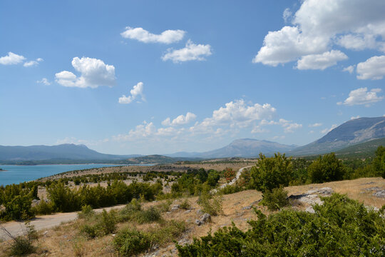 A wide panoramic view from the eastern shore of Peruca Lake looking north toward the distant peaks of the Dinara mountain range under a blue summer sky