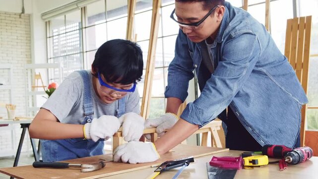 Happy Asian father and son work as a woodworker and carpenter. Father teaching his son to hammer nails on a wooden plank carefully together. carpentry working at a home workshop studio.